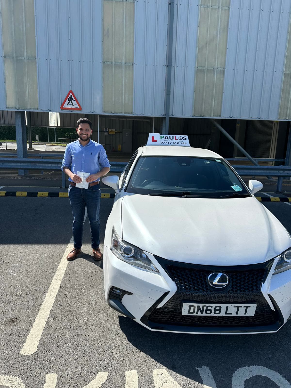 Pupil in a blue shirt holding his driving test pass certificate beside a white Paulos car