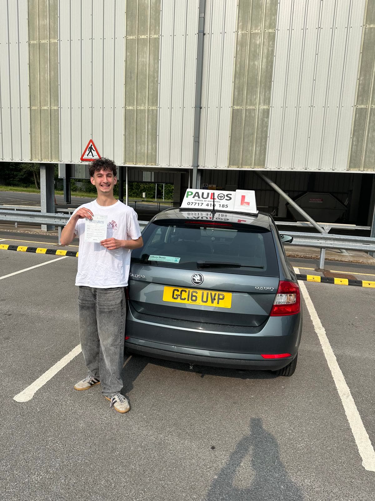 Pupil in a white t-shirt holding his driving test pass certificate beside a Paulos car